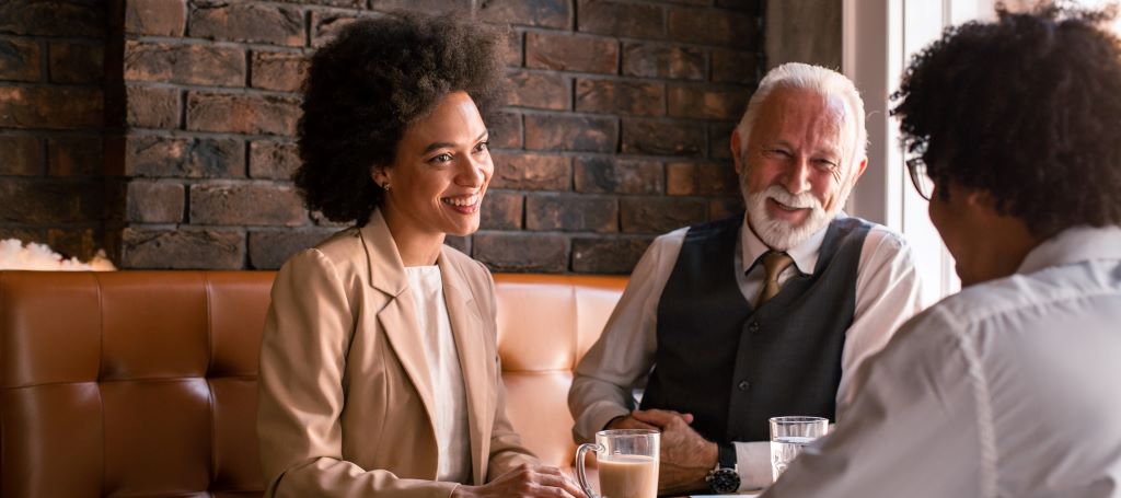 A young BIPOC woman and young BIPOC man laugh over coffee with their older white coworker.