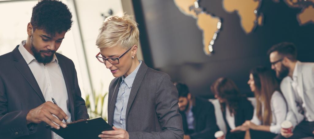 Black businessman and White Businesswoman with look at a clipboard together while colleagues sit in the background.