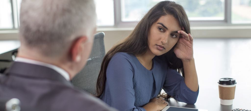 Young South Asian business woman leans on hand and looks seriously at businessman who is talking to her.